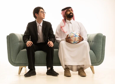 النظر الى شىء ما، بورتريه لرجل عربى خليجى سعودى يرتدى الثوب التقليدى والشماغ  يحمل كورة الفريق يجلس على الاريكة بجانب شاب اسيوى يرتدى البدلة الرسمية، تشجيع فريق كرة قدم، خلفية بيضاء