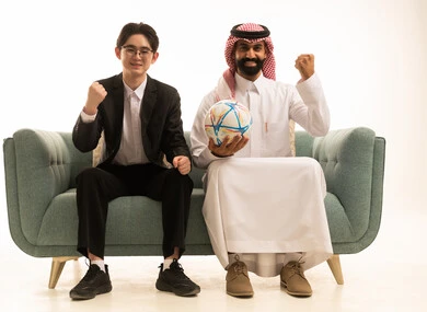 النظر الى الكاميرا بايماءات الفوز و النصر، بورتريه لرجل عربى خليجى سعودى يرتدى الثوب التقليدى والشماغ يرفع يديه بتعابير الفوز و يحمل كورة الفريق يجلس على الاريكة  بجانب شاب اسيوى يرتدى البدلة الرسمية, خلفية بيضاء