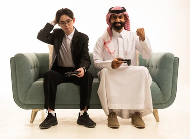 Entertainment and fun with video games in free time, a portrait of a Saudi Gulf Arab man wearing traditional attire and a ghutrah, raising his hands with expressions of victory, sitting on the sofa next to an Asian young man wearing a formal suit, looking at the camera with expressions of regret and sadness, white background.