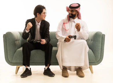 بورتريه لرجل عربى خليجى سعودى يرتدى الثوب التقليدى والشماغ يجلس على الاريكة بجانب شاي اسيوى يرتدى البدلة الرسمية يمسكان عصا التحكم يرفعان يديهما للاعلى بتعبير الفوز, العالم الرقمي الترفيهي, قضاء وقت ممتع بالألعاب الالكترونية، خلفية بيضاء