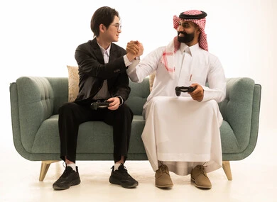 Spending enjoyable time with electronic games, a portrait of a Saudi Gulf Arab man wearing traditional attire and a shemagh sitting on the couch next to a young Asian man in a formal suit, both holding game controllers and raising their hands in victory, the digital entertainment world, white background.