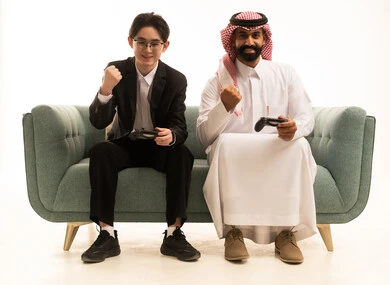 رفع قبضة اليد للاعلى بتعابير الفوز و الانتصار، بورتريه لرجل عربى خليجى سعودى يرتدى الثوب التقليدى والشماغ يجلس على الاريكة بجانب شاب اسيوى يرتدى البدلة الرسمية يمسكان عصا التحكم, العالم الرقمي الترفيهي, خلفية بيضاء