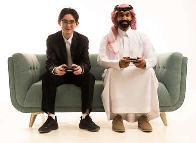 Participation and competition in modern and advanced technical games, a portrait of a Gulf Arab Saudi man wearing traditional attire and a ghutrah sitting on the sofa next to Asian tea, wearing a formal suit holding a game controller, the digital entertainment world, white background.