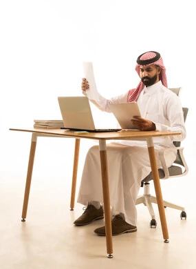 بورتريه لرجل عربى خليجى سعودى يرتدى الثوب التقليدى والشماغ يجلس على مكتب يحمل بيده الاوراق ويقوم بتدقيق العمل امام جهاز الحاسوب المحمول، مفهوم ادارة العمل عن بعد، استخدام التكنولوجيا الحديثة، خلفية بيضاء