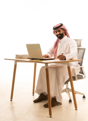 كتابة تقرير العمل، بورتريه لرجل عربى خليجى سعودى يرتدى الثوب التقليدى والشماغ يجلس على مكتب يمسك القلم يقوم بتسجيل ملحوظة امام جهاز الحاسوب المحمول، مفهوم ادارة العمل عن بعد، استخدام التكنولوجيا الحديثة، خلفية بيضاء