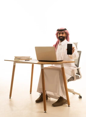 بورتريه لرجل عربى خليجى سعودى يرتدى الثوب التقليدى والشماغ يجلس على مكتب يمسك الهاتف المحمول امام الحاسوب، دمج العمل بالتقنية والتكنولوجيا، عرض شاشة سوداء عبر الجوال، خلفية بيضاء