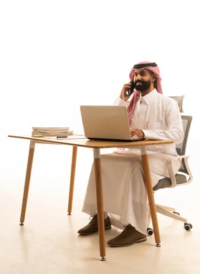 Making a phone call via a mobile phone, a portrait of a Saudi Gulf Arab man wearing traditional attire and a ghutrah sitting at a desk using a computer, merging work with technology and tech, white background.