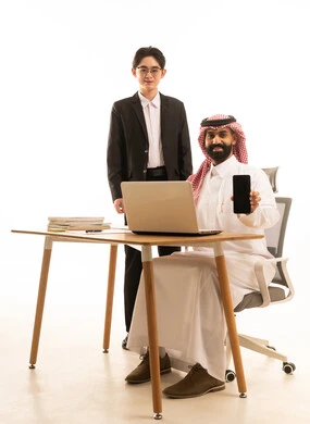 A black screen is displayed on the mobile, a portrait of a Saudi Gulf Arab man wearing traditional attire and a ghutrah sitting at a desk using a mobile phone, next to him stands a young Asian man in formal attire, merging work with technology and tech, white background.