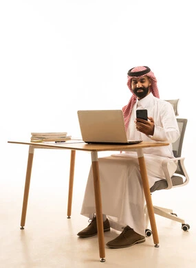 اجتماع عمل أونلاين، بورتريه لرجل عربى خليجى سعودى يرتدى الثوب التقليدى والشماغ يجلس على مكتب يستخدم الهاتف المحمول، استخدام التقنية الحديثة، خلفية بيضاء