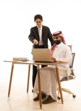 دمج العمل بالتقنية والتكنولوجيا، الاشارة الى الحاسوب، بورتريه لرجل عربى خليجى سعودى يرتدى الثوب التقليدى والشماغ يجلس على مكتب يستخدم الحاسوب يقف بجانبه شاب اسيوى يرتدي البدلة الرسمية يتعلمان اساليب العمل والتطوير المهني، خلفية بيضاء