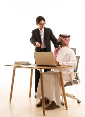 The reference to the computer, integrating work with technology and tech, a portrait of a Gulf Arab Saudi man wearing traditional attire and a shemagh sitting at a desk using a computer, next to him stands an Asian young man in a formal suit, both learning work methods and professional development, with a white background.