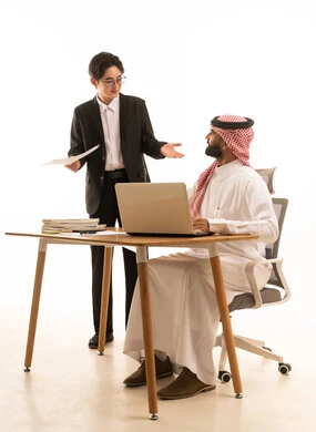 تطوير العمل وحل المشكلات، بورتريه لرجل عربى خليجى سعودى يرتدى الثوب التقليدى والشماغ يجلس على مكتب يستخدم الحاسوب يقف بجانبه شاب اسيوى يرتدي البدلة الرسمية يتعلمان اساليب العمل والتطوير المهني، خلفية بيضاء
