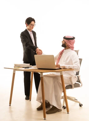 Greetings and peace, and the agreement on a successful business deal. A portrait of a Saudi Gulf Arab man wearing traditional attire and a ghutrah sitting at a desk using a computer, next to him stands a young Asian man in a formal suit. They are learning work methods and professional development, with a white background.