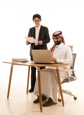 العمل الجماعى و روح الفريق الواحد، بورتريه لرجل عربى خليجى سعودى يرتدى الثوب التقليدى والشماغ يجلس على مكتب يستخدم الحاسوب يقف بجانبه شاب اسيوى يرتدي البدلة الرسمية يتعلمان اساليب العمل والتطوير المهني، استخدام جهاز اللابتوب،  خلفية بيضاء