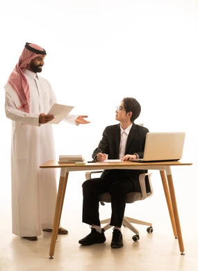 استخدام جهاز الحاسوب، وضع الخطة الاستراتيجية للعمل، بورتريه لرجل عربى خليجى سعودى يرتدى الثوب التقليدى والشماغ يتناقش مع شاب اسيوى يرتدي البدلة الرسمية و يتعلمان اساليب العمل والتطوير المهني، روح الفريق الواحد والعمل الجماعي، خلفية بيضاء
