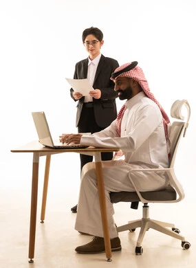 بورتريه لرجل عربى خليجى سعودى يرتدى الثوب التقليدى والشماغ يجلس على مكتب يستخدم الحاسوب يقف بجانبه شاب اسيوى يرتدي البدلة الرسمية يتعلمان اساليب العمل والتطوير المهني، روح الفريق الواحد، استخدام جهاز اللابتوب،  خلفية بيضاء