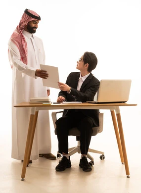 Develop a strategic work plan, a portrait of a Saudi Gulf Arab man holding a project report discussing with a young Asian man in a formal suit sitting behind a desk using a computer to complete work tasks, studying and analyzing projects, white background.
