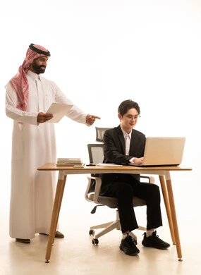 بورتريه لرجل عربى خليجى سعودى يمسك تقرير مشروع يقف بجانب شاب اسيوى يرتدي البدلة الرسمية يجلس خلف طاولة المكتب يستخدم الحاسوب لانجاز مهام العمل،  دراسة وتحليل المشاريع، خلفية بيضاء