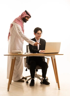 الشراكة والتعاون في إنجاز المهام، بورتريه لشاب اسيوى يرتدي البدلة الرسمية يجلس خلف طاولة المكتب يتناقش مع رجل عربى خليجى سعودى فى اجتماع عمل، دراسة وتحليل المشروع باستخدام الحاسوب، خلفية بيضاء