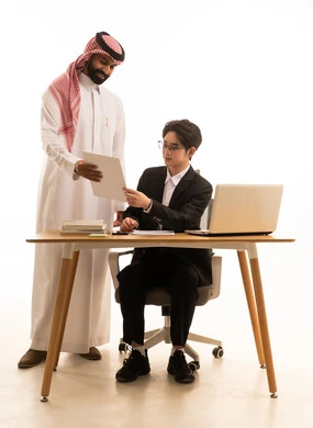 تبادل الخبرة والمعلومة لتحقيق هدف الشركة، بورتريه لرجل عربى خليجى سعودى يمسك تقرير مشروع يتناقش مع شاب اسيوى يرتدي البدلة الرسمية يجلس خلف طاولة المكتب يستخدم الحاسوب لانجاز مهام العمل، دراسة وتحليل المشاريع، خلفية بيضاء