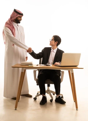 بورتريه لشاب اسيوى يرتدى البدلة الرسمية يجلس على  طاولة المكتب يصافح بيده رجل عربى خليجى سعودى يترتدى الثوب التقليدى والشماغ، الترحيب و القاء السلام، استخدام جهاز تقني حديث و متطورلانجاز المشروع، العمل عن بعد،  خلفية بيضاء