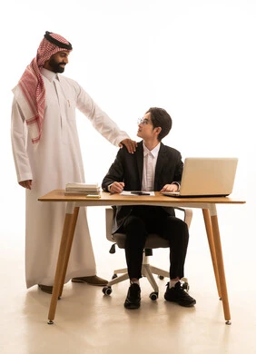 روح الفريق الواحد والعمل الجماعي، استخدام جهاز الحاسوب، وضع الخطة الاستراتيجية للعمل، بورتريه لرجل عربى خليجى سعودى يرتدى الثوب التقليدى والشماغ يتناقش مع شاب اسيوى يرتدي البدلة الرسمية و يتعلمان اساليب العمل والتطوير المهني، خلفية بيضاء