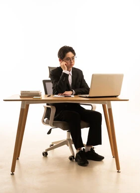 محاولة تذكر أمر ما، استخدام جهاز تقنى حديث، بورتريه لشاب اسيوى يرتدي البدلة الرسمية  يجلس خلف طاولة خشبية امام اللابتوب يضع يده على رأسه بتعابير الحيرة، مفهوم ادارة العمل عن بعد، خلفية بيضاء