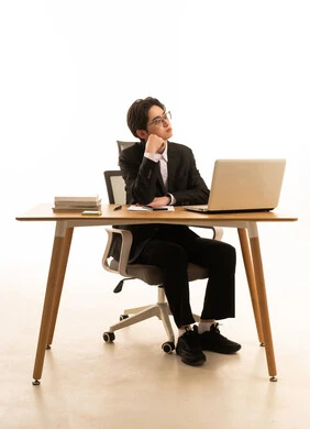 بورتريه لشاب اسيوى يرتدي البدلة الرسمية  يجلس خلف طاولة خشبية امام اللابتوب يضع يده على خده بتعابير الحيرة، محاولة تذكر أمر ما، استخدام جهاز تقنى حديث، مفهوم ادارة العمل عن بعد، خلفية بيضاء