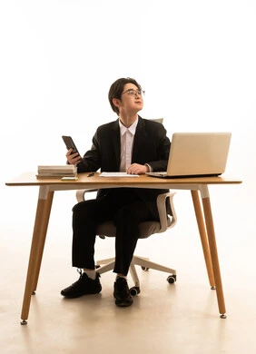 النظر الى شىء ما، استخدام جهاز تقني حديث و متطورلانجاز المشروع، العمل عن بعد، بورتريه لشاب اسيوى يرتدى البدلة الرسمية يجلس على  طاولة المكتب يستخدم الهاتف المحمول امام جهاز الحاسوب، خلفية بيضاء