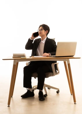 Making a phone call via mobile, using a laptop to complete tasks, a portrait of a young Asian man in a formal suit sitting behind a wooden table using a laptop and a phone, the concept of remote work management, white background.