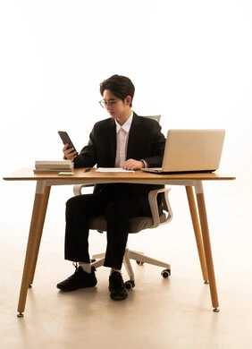 تنظيم وادارة جدول مهام العمل، استخدام جهاز تقنى حديث، بورتريه لشاب اسيوى يرتدي البدلة الرسمية  يجلس خلف طاولة خشبية امام اللابتوب يمسك الهاتف المحمول، مفهوم ادارة العمل عن بعد، خلفية بيضاء