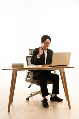 Raising the hand to greet, using a laptop to accomplish tasks, a portrait of a young Asian man in a formal suit sitting behind a wooden table in front of the laptop making a video call via the laptop, the concept of remote work management, white background.