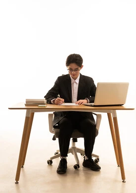 كتابة و تدوين الملاحظات، إستخدام جهاز الحاسوب المحمول لإنجاز المهام، بورتريه لشاب اسيوى يرتدي البدلة الرسمية  يجلس خلف طاولة خشبية امام اللابتوب يقوم بكتابة تقرير، مفهوم ادارة العمل عن بعد، خلفية بيضاء