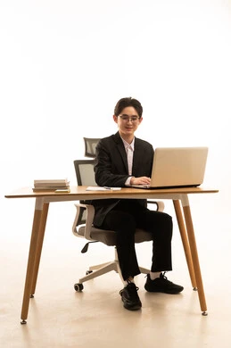 Looking at the camera with gestures of joy, using a laptop to accomplish tasks, a portrait of a young Asian man in a formal suit sitting behind a wooden table using a laptop looking at something, the concept of remote work management, white background.