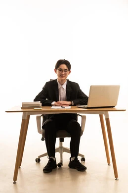 A portrait of a young Asian man wearing a formal suit sitting behind a wooden table using a computer and looking at the camera, the concept of remote work management, the use of modern devices and advanced technologies, white background.