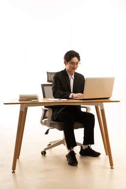 مفهوم ادارة العمل عن بعد، بورتريه لشاب اسيوى يرتدي البدلة الرسمية  يجلس خلف طاولة خشبية يستخدم الحاسوب، استخدام الأجهزة الحديثة والتقنيات المتطورة، خلفية بيضاء