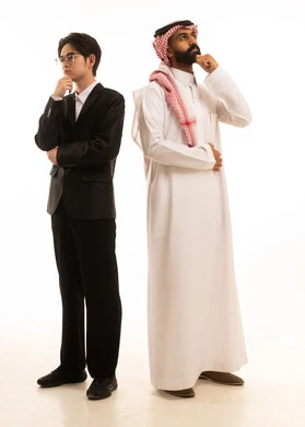 التمعن والتركيز لإيجاد الحل لمشكلة ما, الشعور بالضيق والملل، بورتريه لرجل عربي خليجي سعودى يرتدي الثوب التقليدى والشماغ يقف و ظهره الى ظهر شاب اسيوى يرتدي البدلة الرسمية يضعان يديهما على وجههما بإيماءات التفكير، الجسم بالطول الكامل، خلفية بيضاء