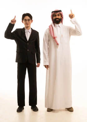 الوقوف باستقامة ورفع اصبعي السبابة في كلتا اليدين، بورتريه لرجل عربي خليجي سعودى يرتدي الثوب التقليدى والشماغ يقف بجانب شاب اسيوى يرتدي البدلة الرسمية يقومان بالاشارة الى مكان ما، الجسم بالطول الكامل، خلفية بيضاء