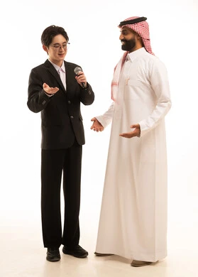 Conducting an interview and asking questions, a portrait of a young Asian man in a formal suit holding a microphone speaking to a Saudi Arabian Gulf man wearing traditional attire and a ghutrah, effective communication and exchange of dialogues and discussions, full-body image, white background.