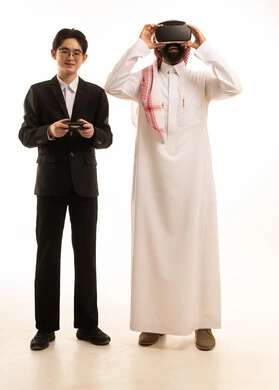 Entertainment through viewing via virtual reality glasses, a portrait of a Saudi Arabian Gulf man wearing traditional attire and a ghutrah looking through 3D glasses standing next to a young Asian man in a formal suit holding a joystick, full-body, white background.