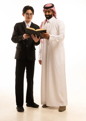 الاستمتاع بقراءة الكتب، قضاء الوقت بالقراءة والتعلم والتثقف، بورتريه لرجل عربي خليجي سعودى يرتدي الثوب التقليدى والشماغ  يقف بجانب شاب اسيوى يرتدي البدلة الرسمية  يقومان بمشاركه قراءة كتاب، صورة الجسم بالطول الكامل، خلفية بيضاء