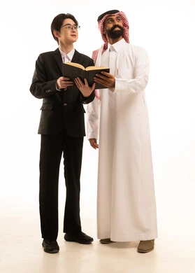 قضاء الوقت بالقراءة والتعلم والتثقف، بورتريه لرجل عربي خليجي سعودى يرتدي الثوب التقليدى والشماغ  يقف بجانب شاب اسيوى يرتدي البدلة الرسمية  يقومان بمشاركه قراءة كتاب و ينظران الى شىء ما، صورة الجسم بالطول الكامل، خلفية بيضاء