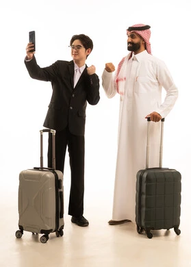 A portrait of a Saudi Arabian Gulf man wearing traditional attire and a shemagh standing next to an Asian young man in a formal suit, both holding travel bags, taking pictures with victory gestures, capturing a selfie using a mobile phone, full-body image, white background.