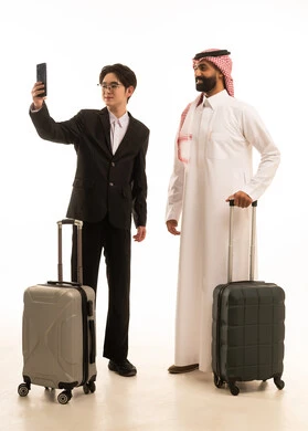 Taking a selfie using a mobile phone, preparing for travel and vacation, a portrait of a Saudi Arabian Gulf man wearing traditional attire and a shemagh standing next to a young Asian man in a formal suit, each holding a travel bag, full-body image, white background.