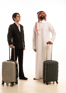 Looking at a place, having a good time on vacation, a portrait of a Saudi Arabian Gulf man wearing traditional attire and a ghutrah standing next to an Asian young man in a formal suit, both holding travel bags and looking at something, full-body image, white background.