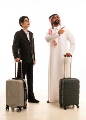 Pointing to a place, having a good time on vacation, a portrait of a Saudi Arabian Gulf man wearing traditional attire and a ghutrah standing next to an Asian young man in a formal suit, both holding a travel bag and looking at something, full-body image, white background.