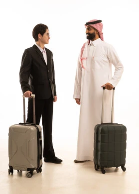 قضاء وقت ممتع في الإجازة، بورتريه لرجل عربي خليجي سعودى يرتدي الثوب التقليدى والشماغ يقف بجانب شاب اسيوى يرتدي البدلة الرسمية يحمل كل منهما حقيبة السفر، الاستمتاع بالسفر و الرحلة الصيفية، صورة الجسم بالطول الكامل، خلفية بيضاء