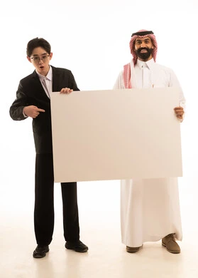 بورتريه لرجل عربي خليجي سعودى يرتدي الثوب التقليدى والشماغ يقف بجانب شاب اسيوى يرتدي البدلة الرسمية يحملان موك اب مستطيل كبير، الاشارة الى لوحة توضيحية بيضاء ،صورة الجسم بالطول الكامل، خلفية بيضاء