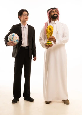 النظر الى شىء ما، تشجيع الفريق السعودي، بورتريه لرجل عربي خليجي سعودى يرتدي الثوب التقليدى و الشماغ يحمل الكأس يقف بجانب شاب اسيوى يرتدي البدلة الرسمية يحمل كورة القدم و يرفع يديه بتعابير الانتصار، الجسم بالطول الكامل، خلفية بيضاء