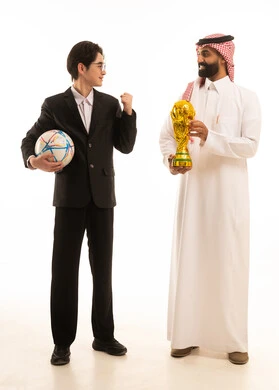 الشعور بالفرح والسرور، تشجيع الفريق السعودي، بورتريه لرجل عربي خليجي سعودى يرتدي الثوب التقليدى و الشماغ يحمل الكأس يقف بجانب شاب اسيوى يرتدي البدلة الرسمية يحمل كورة القدم و يرفع يديه بتعابير الانتصار، الجسم بالطول الكامل، خلفية بيضاء